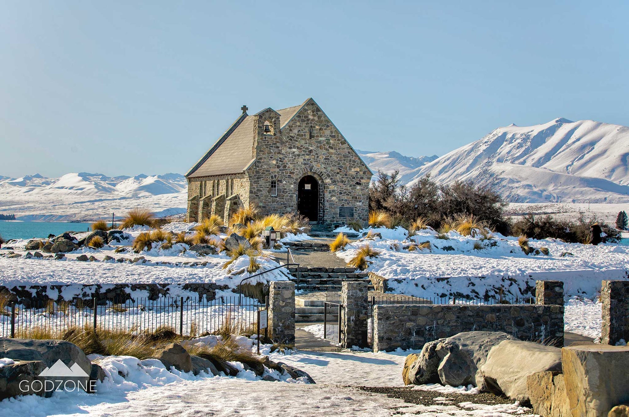 Photograph of stone church at Lake Tekapo in snow with snowy mountains. White, grey and pale blue tones. For sale.