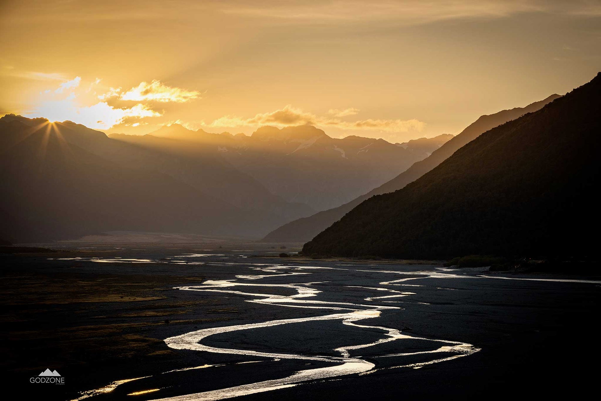 Landscape photograph of the sun setting over a braided river in NZ South Island's Arthurs Pass. Light reflecting off the water. Evening glow.