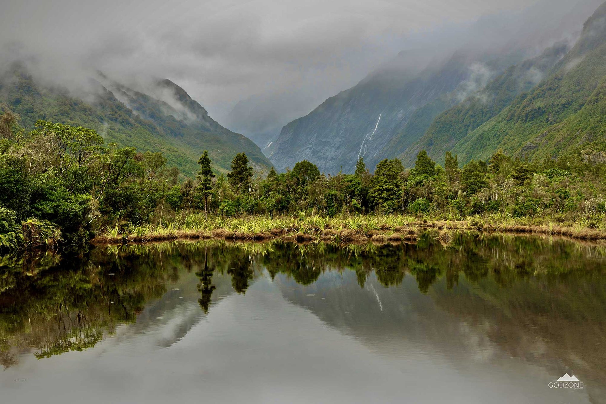 Atmospheric landscape photograph of peters pool in New Zealand's South Island with misty mountains, waterfalls, green bush and shrub.