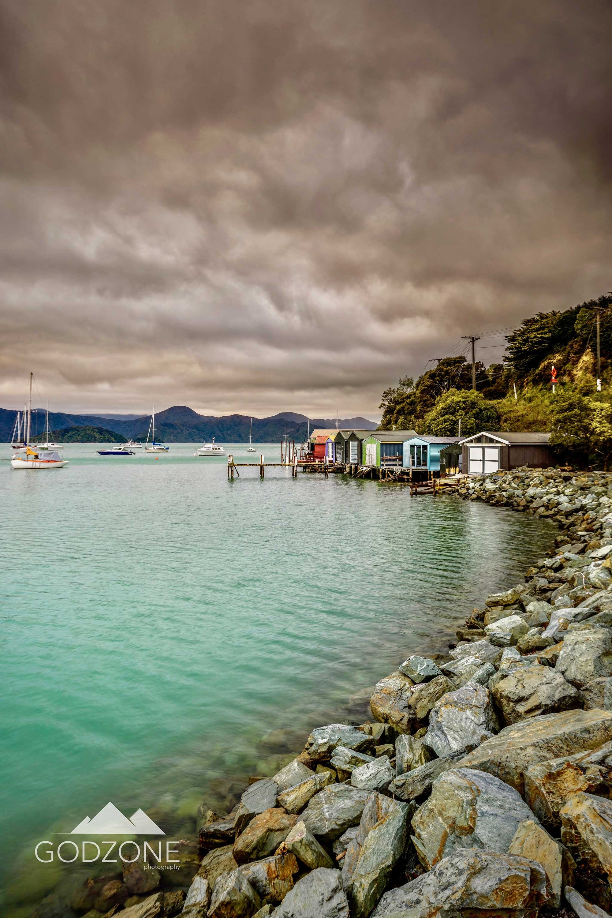 Waikawa Bay photograph with stony bank, coloured boat houses, and stormy skies above. Beautifully captured and composed photograph for sale.