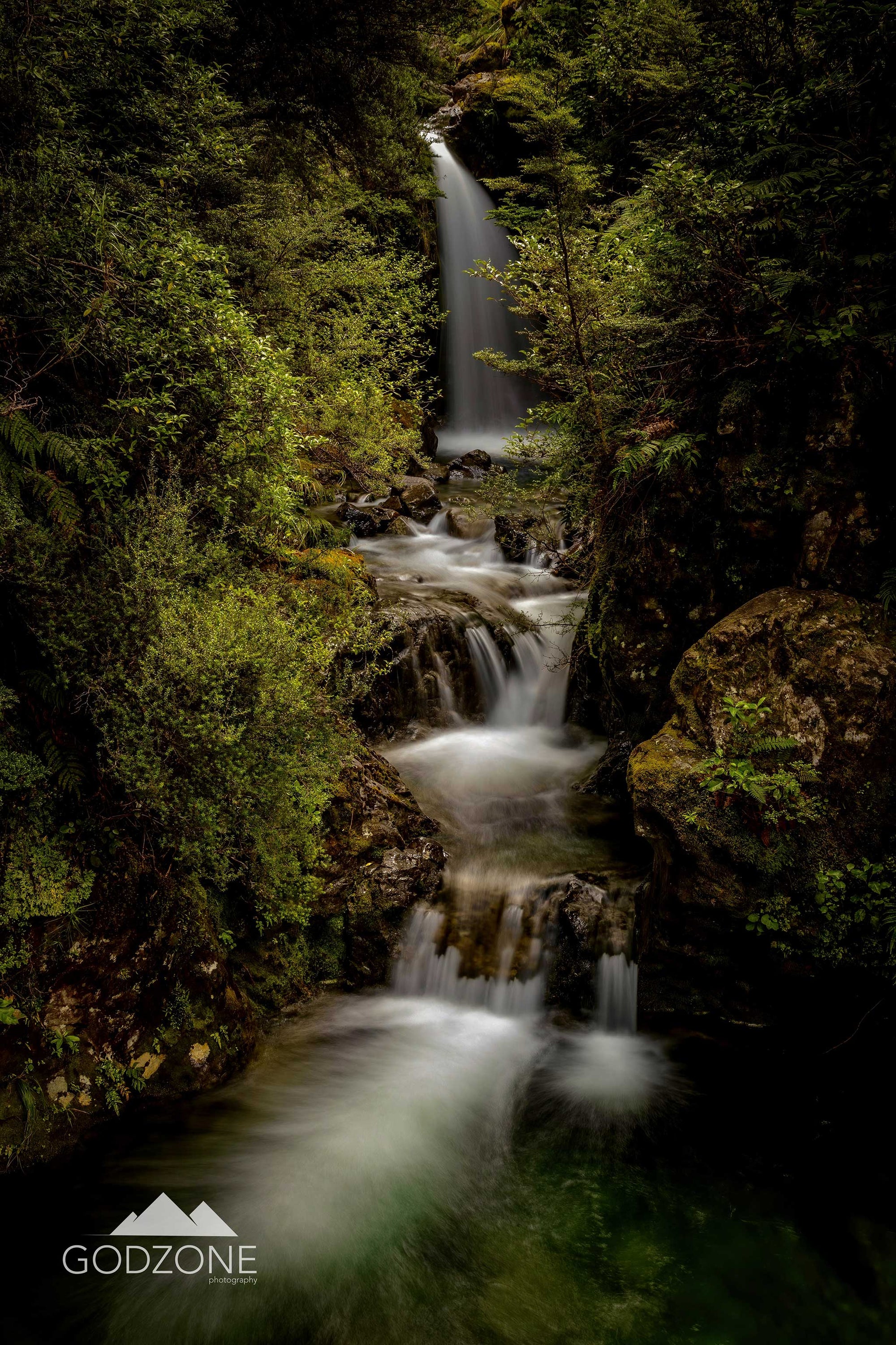 Long-exposure portrait photograph of the Avalanche Falls in Arthur's Pass, South Island, New Zealand. Green bush and soft white falling water.