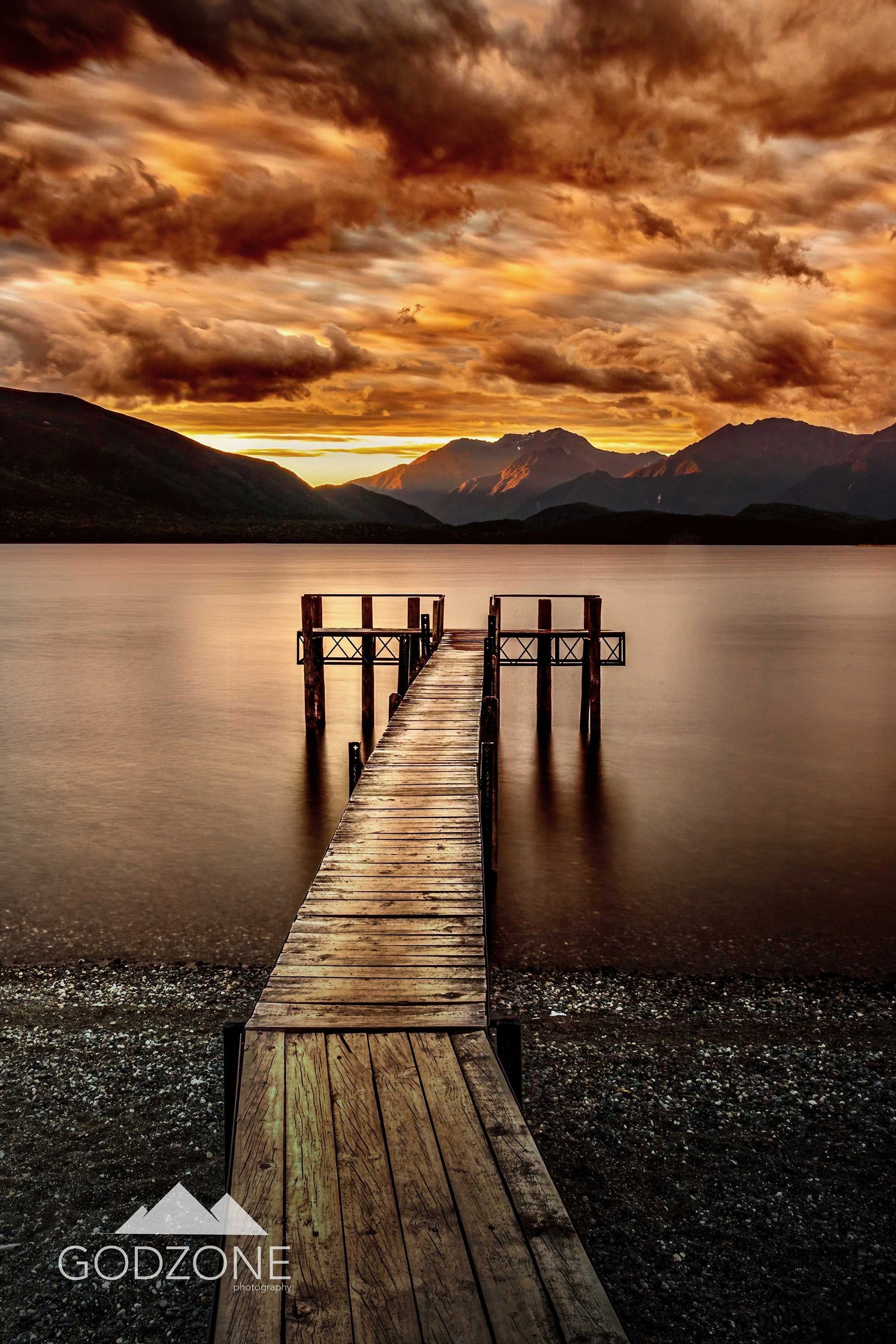 Stunning tall portrait photograph of stormy orange skies and a sunset over the jetty at Te Anau in NZ's south island. Beautiful golden light on the mountains.