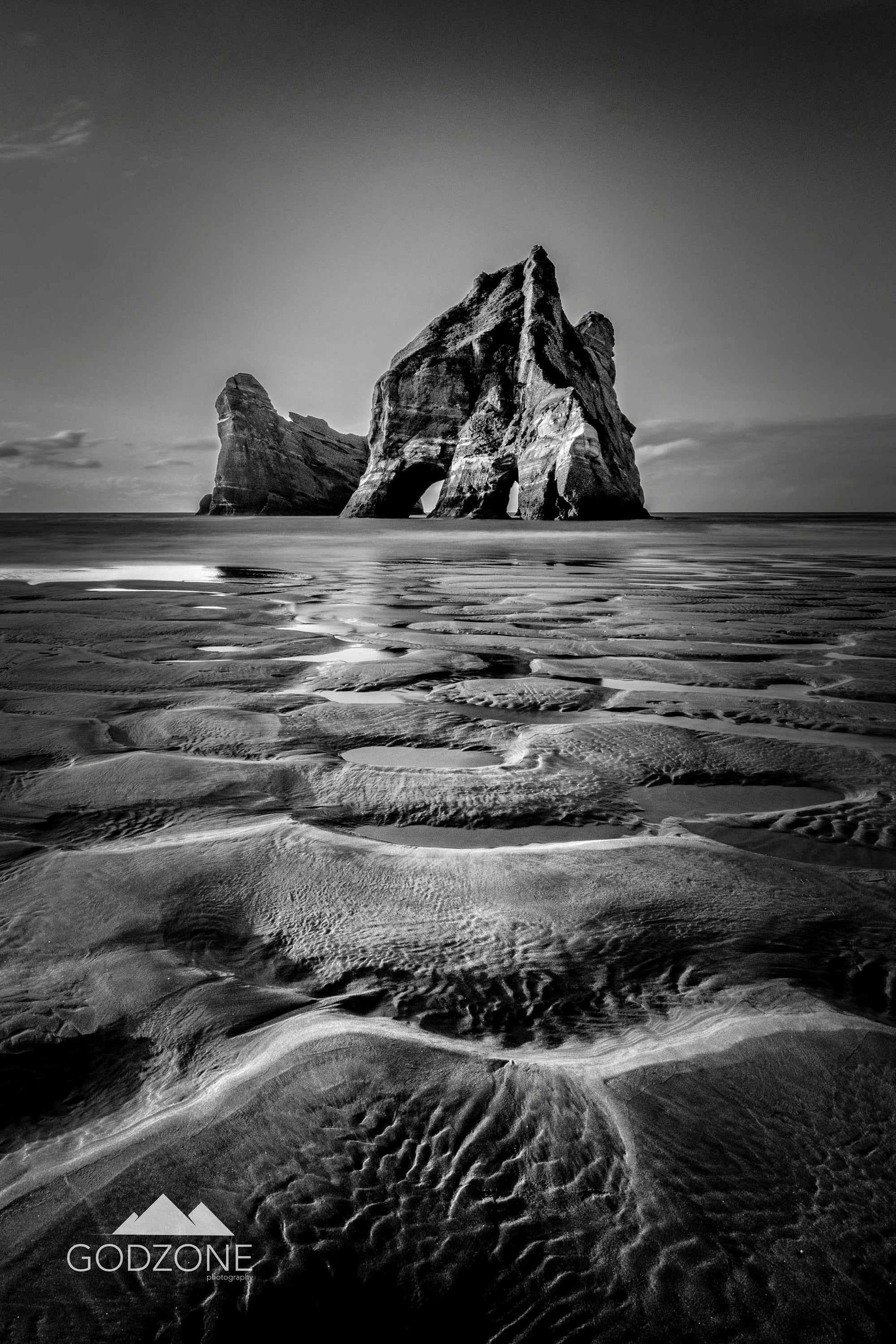 Black and white portrait photograph of rippled sand leading to archway islands in NZ South Island.