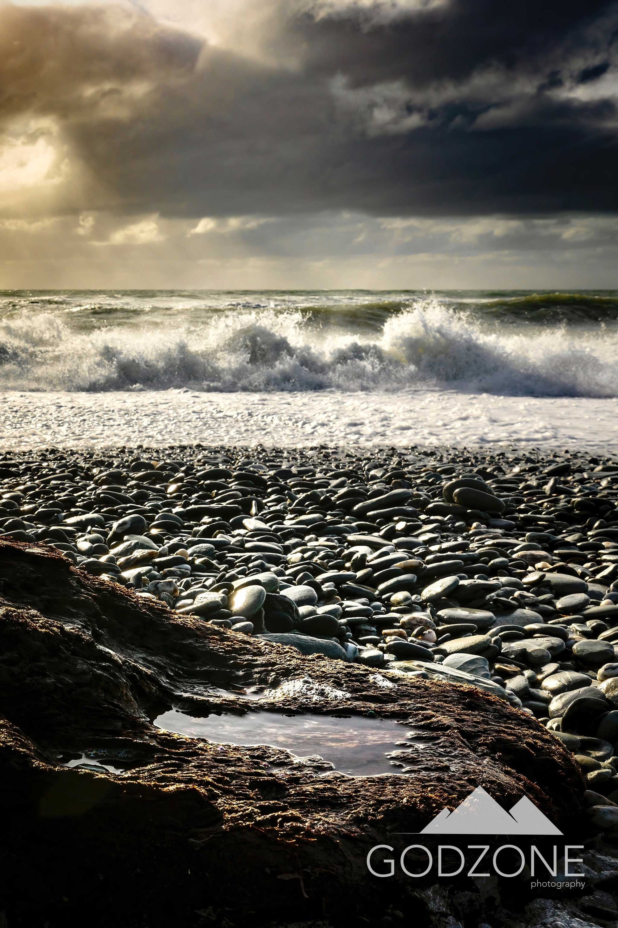 Rugged West Coast beach photograph with pebbles, crashing waves, soft light through stormy skies. NZ tall landscape photograph.