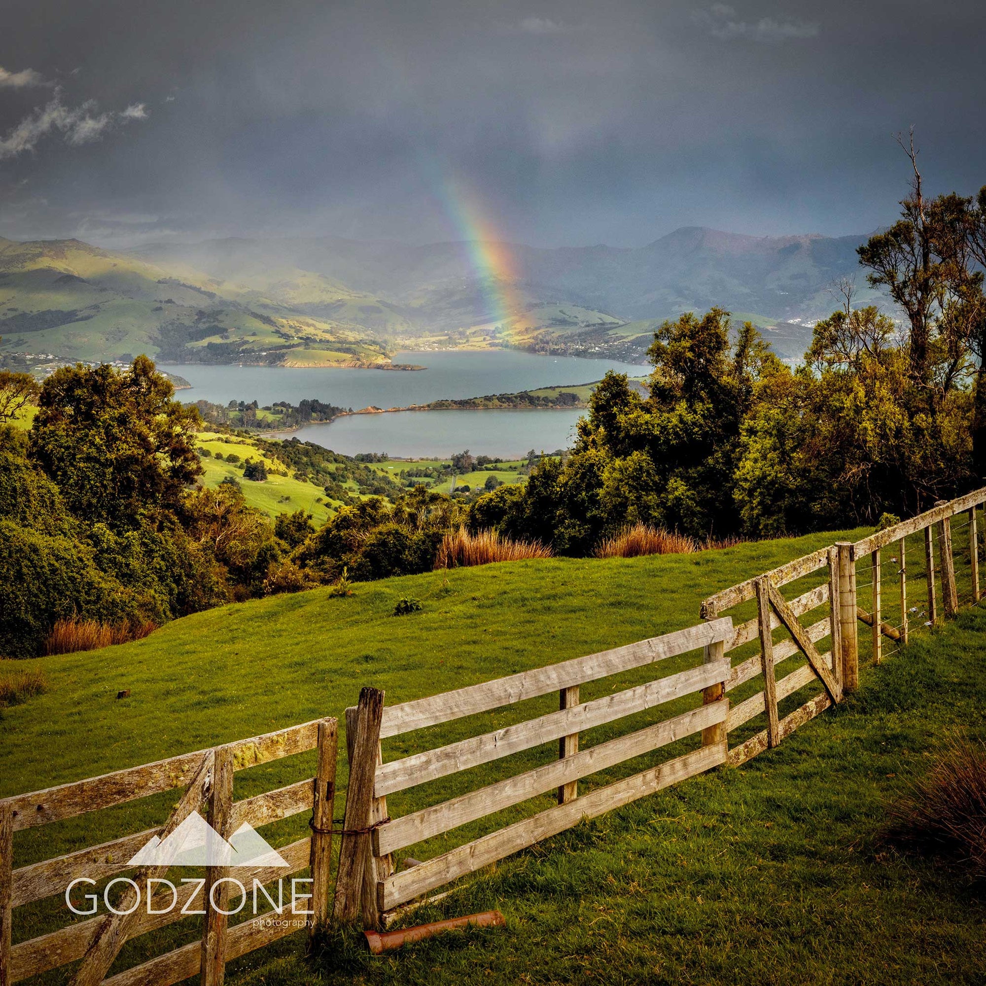 Square photograph of banks peninsula with a fence and looking down on a small rainbow forming over the water. South Island, New Zealand.