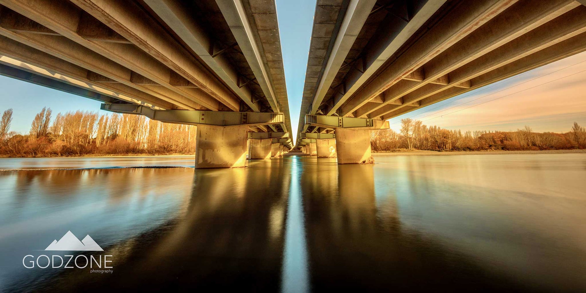 Striking landscape photograph of the Waimakariri bridges seen from below. Golden and blue colour tones. 