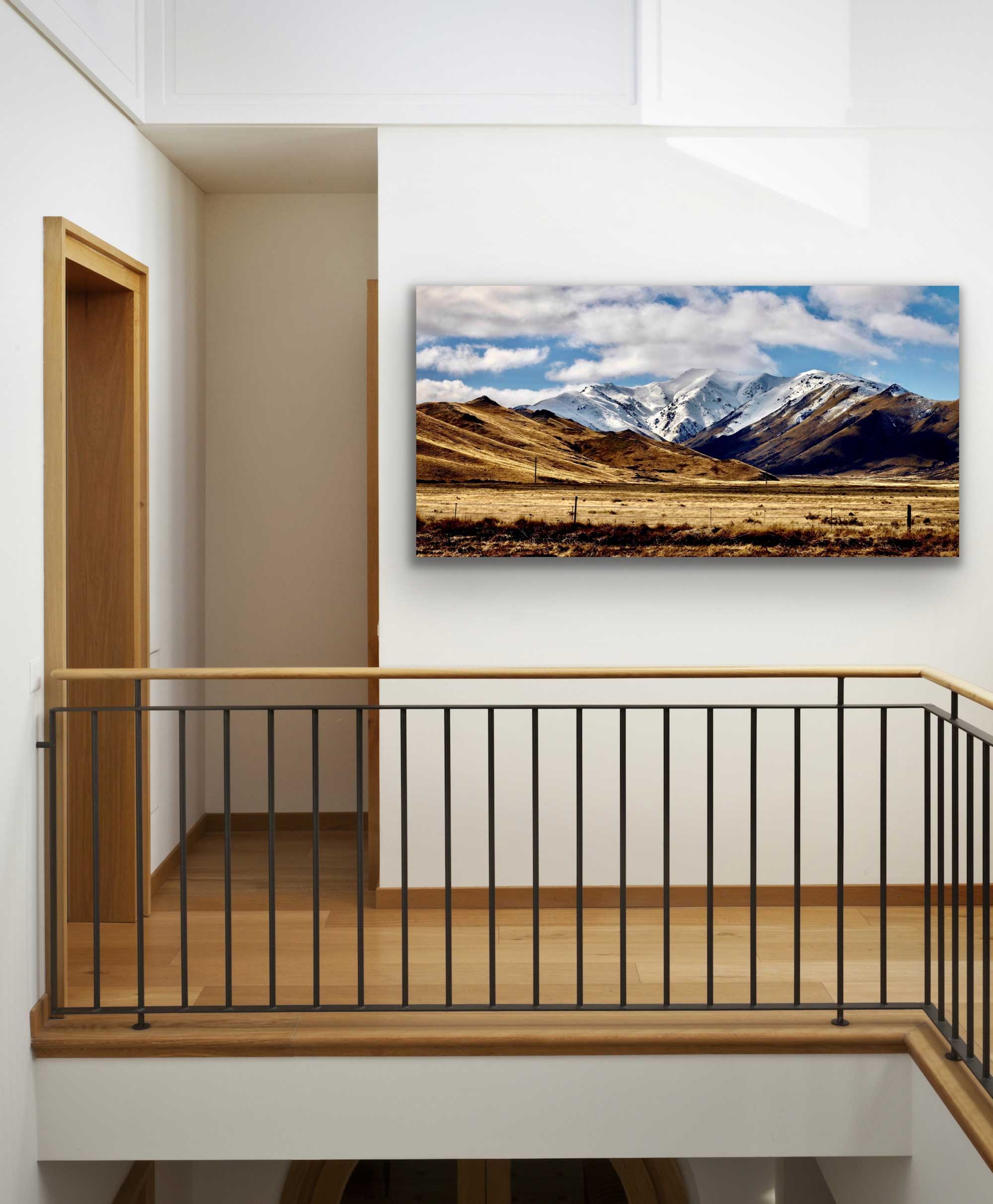 In situ wide, panoramic landscape photograph of Burkes mountain pass in New Zealand's South Island. Snowy mountains and golden fields. Photograph on wall above stairway.