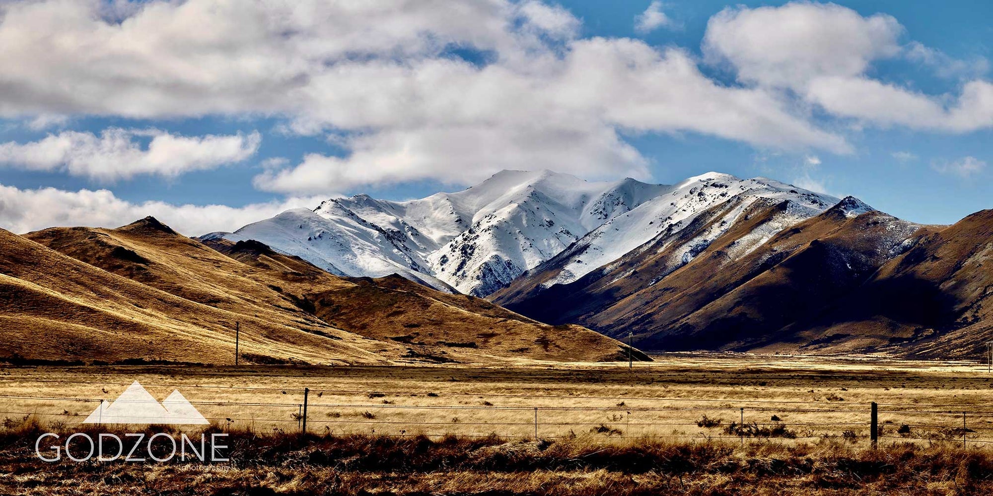 Wide, panoramic landscape photograph of Burkes mountain pass in New Zealand's South Island. Snowy mountains and golden fields.