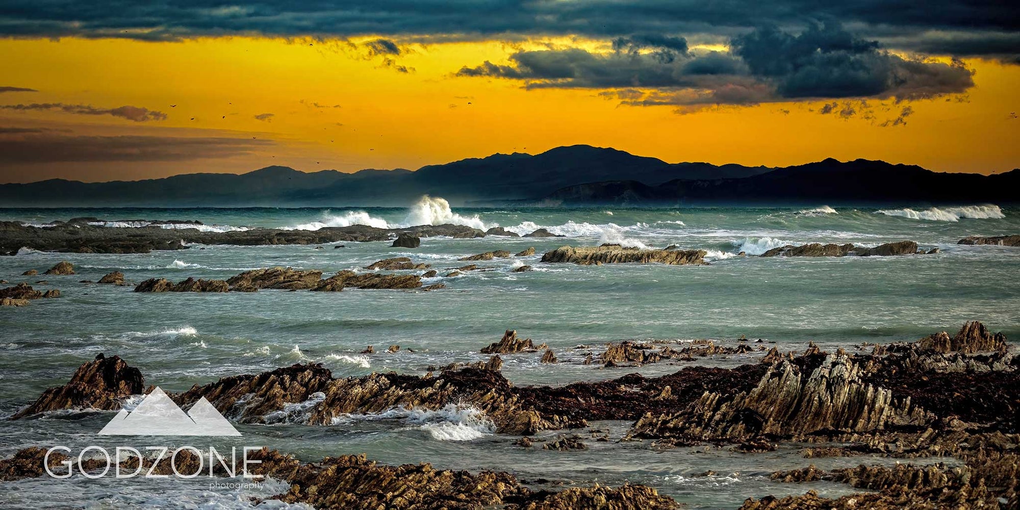 NZ landscape photograph of crashing waves and a yellow sunset at South Bay, Kaikoura. Rugged rocky coastline. NZ photographs for sale.