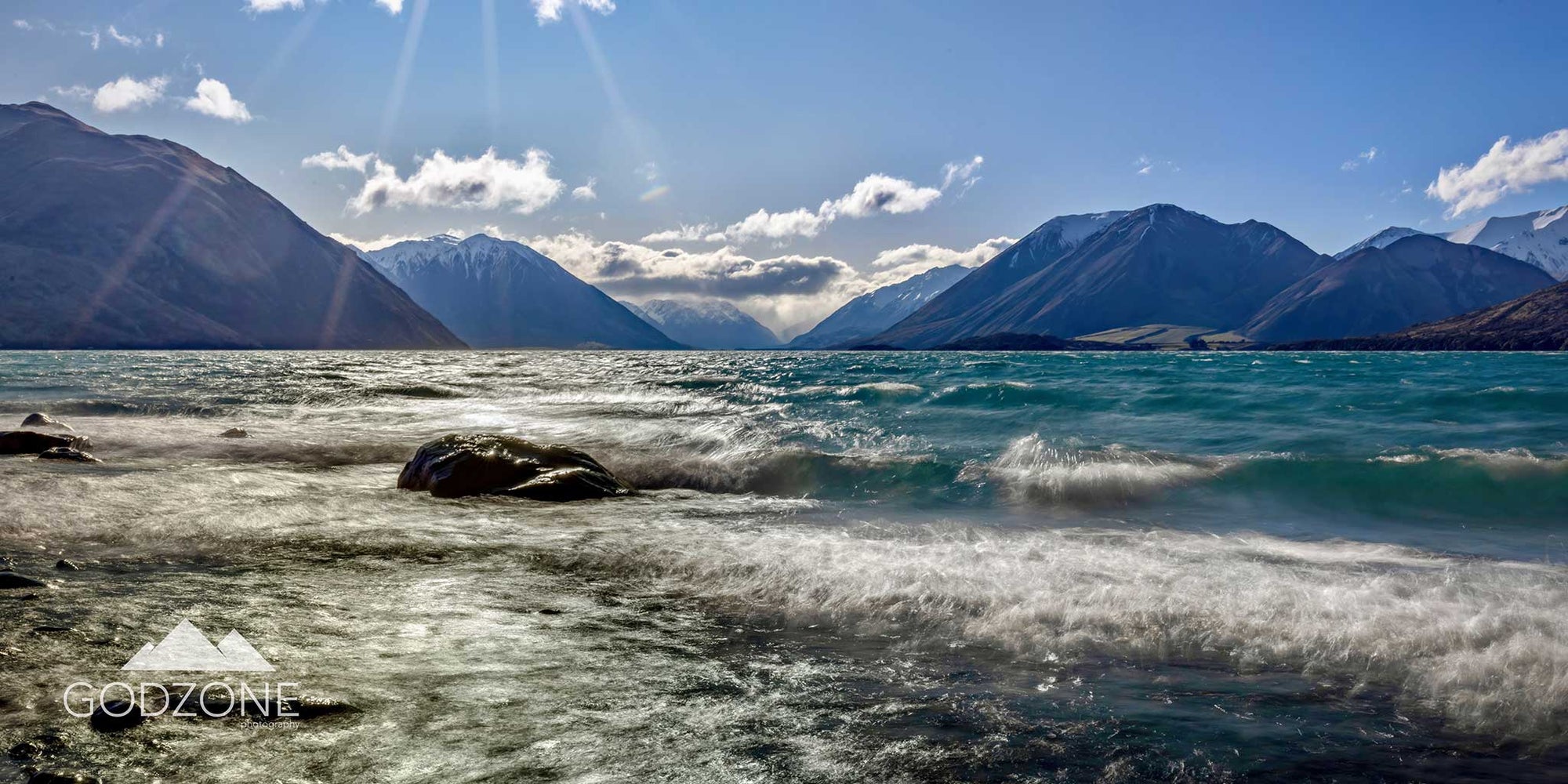 Bright and blue South Island landscape photograph of Lake Coleridge with sun rays and misty blue snow-tipped mountains.