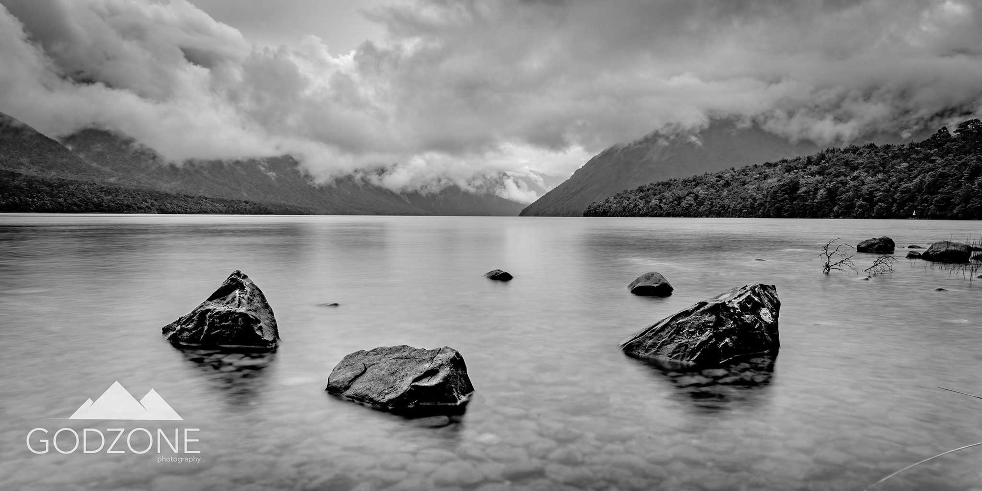 Subtle black and white or greyscale photograph of Lake Rotoiti with large stone in high detail and pebbles perfectly caught beneath the water. 