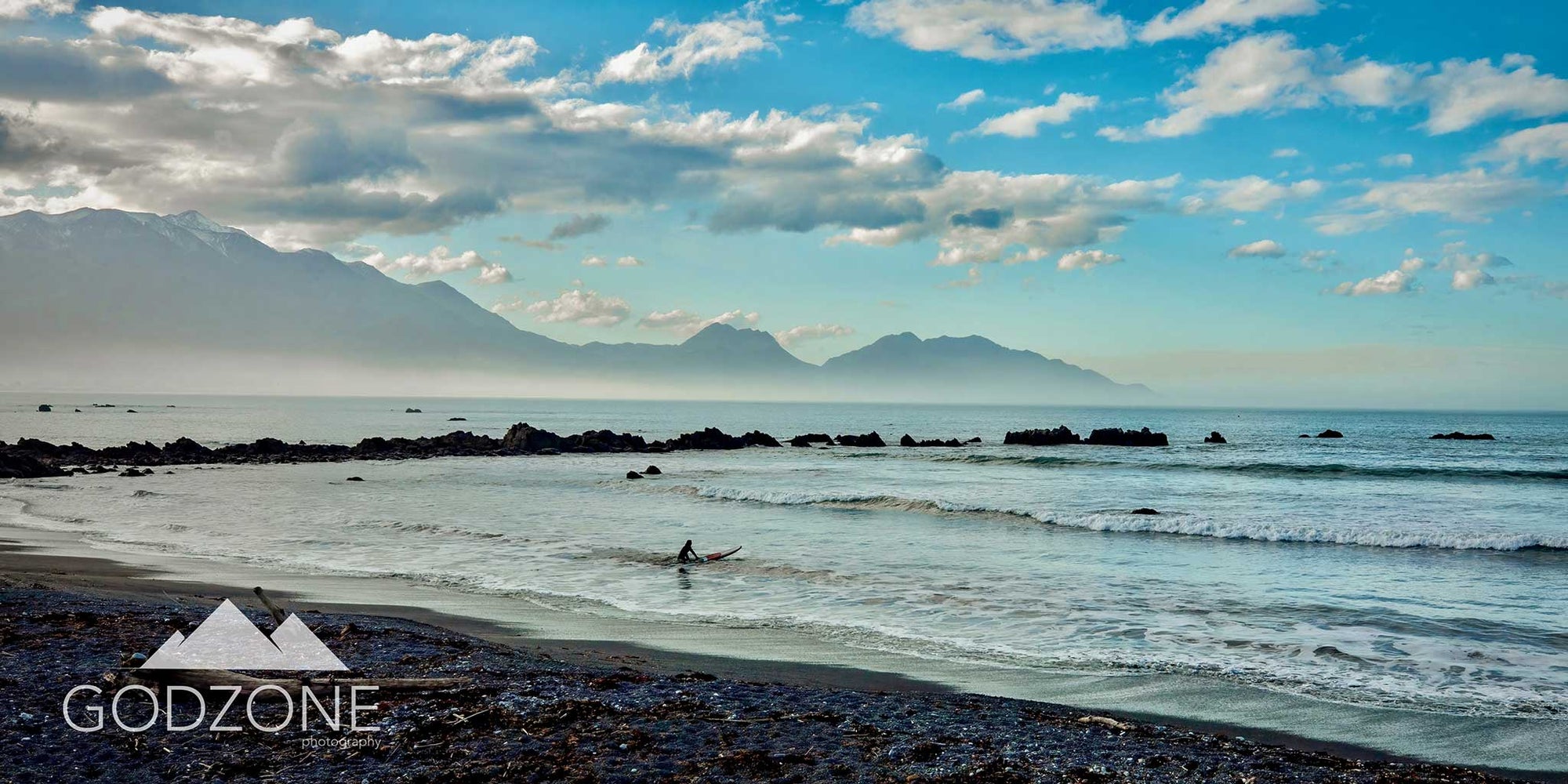 Beautiful panoramic landscape photograph of a surfer in blue waves and skies at Kaikoura beach in New Zealand's South Island. Blue and grey tone artwork for sale. Surfing photography NZ.