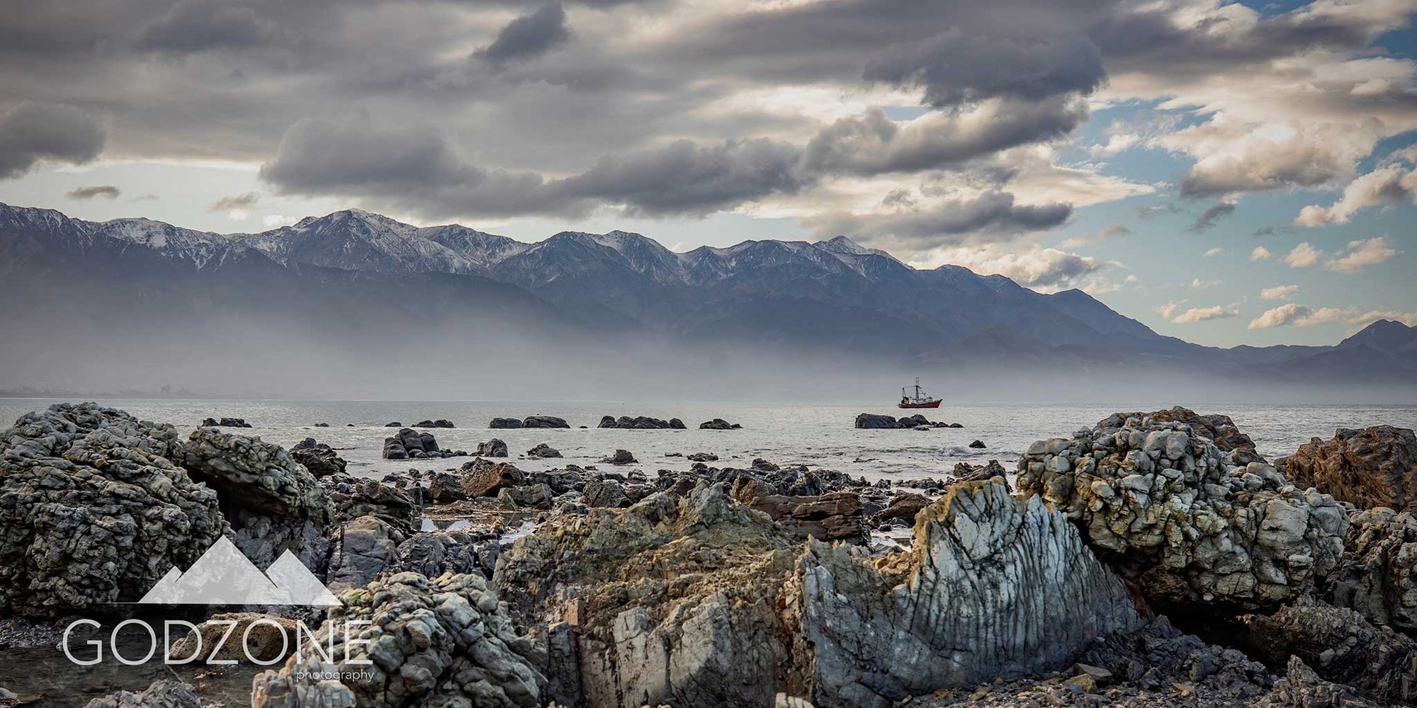 Rugged subtle colours landscape photograph of rocky shoreline, boat, light shafting through mountains in Kaikoura, North Canterbury. NZ South Island landscape photography. Grey tone artwork.